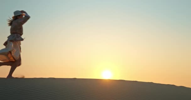 Feliz mulher animada correndo por duna de areia com pôr do sol dourado no fundo, 4K — Vídeo de Stock