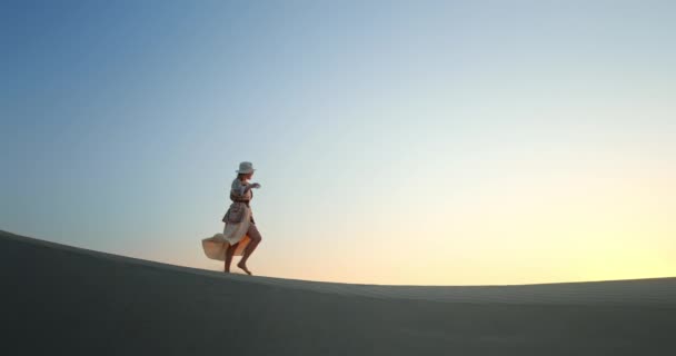 Mujer feliz caminando en el desierto al atardecer. Chica positiva lanzando sombrero de paja en el aire — Vídeos de Stock