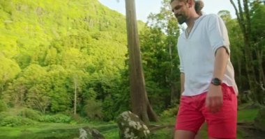 Sakallı ve at kuyruklu bir adam yeşilliklerle çevrili gölete doğru yürüyor.