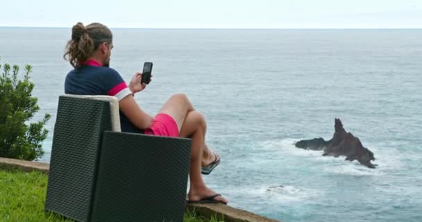 Άνδρας με μαύρο t-shirt και φούξια σορτς διαβάζει από μια τηλεφωνική οθόνη — Αρχείο Βίντεο