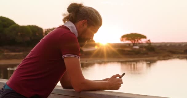 Caucasien mec en rouge t-shirt textos avec coucher de soleil ciel en arrière-plan — Video