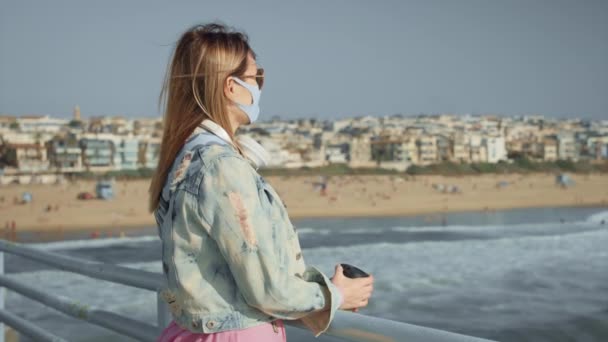 Mulher bonita em máscara protetora no cais do oceano com vista para a cidade da praia 8K — Vídeo de Stock