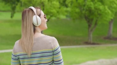 Positive young woman listening music in wireless white headphones in green park