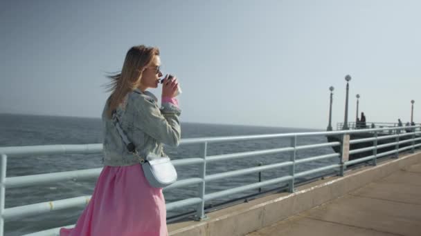 Стильна щаслива жінка в рожевій сукні, що йде океанським пірсом, п'є каву — стокове відео
