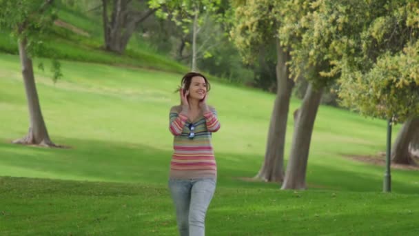 Happy excited smiling young woman jumping while listening music in green park — Stockvideo