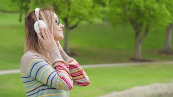 Young woman relaxing in green park on summer day in wireless white headphones 8K — Stok video