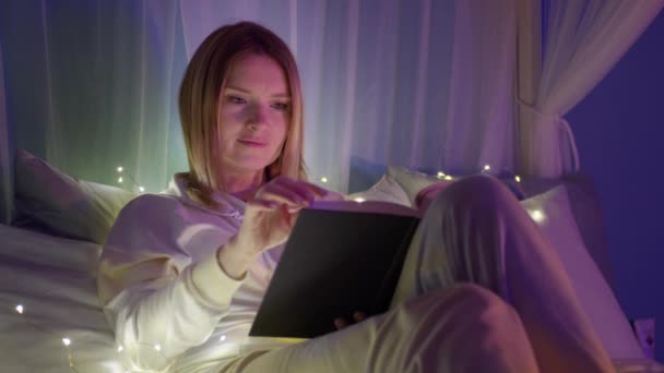 Спокойная, расслабленная молодая женщина читает книгу с волшебными огнями в уютной домашней атмосфере — стоковое видео