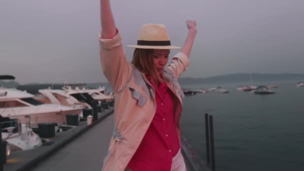 Młoda kobieta w stylowej czapce i modnym płaszczu tańczy na przystani o zachodzie słońca 4K — Wideo stockowe