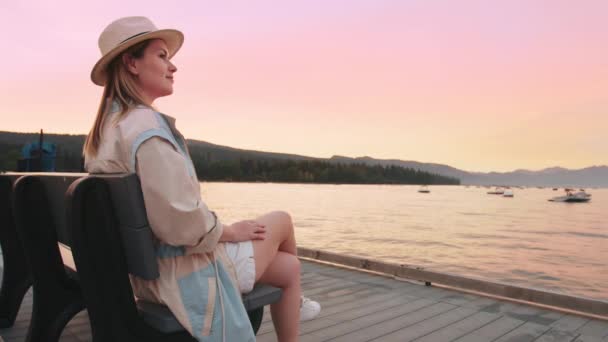Podróżnik siedzi na ławce ciesząc się różowym zachodem słońca odzwierciedlającym w złotych wodach jeziora — Wideo stockowe