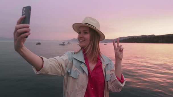 Mujer sonriente feliz usando mascarilla divertida en el teléfono inteligente al aire libre, puesta de sol en el lago — Vídeo de stock