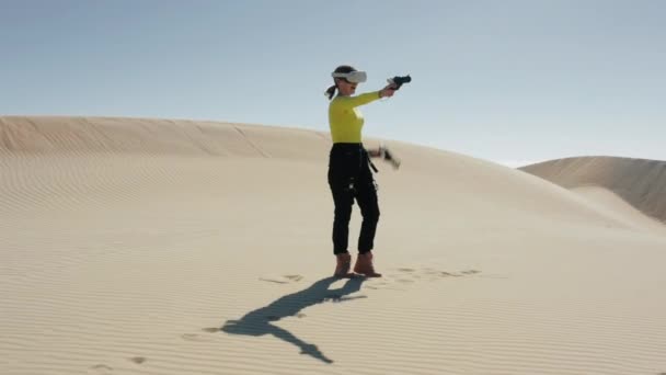 Sanal gerçeklik kulaklığı takmış enerjik genç bir kadın kum tepelerinde VR nişancılığını oynuyor. — Stok video