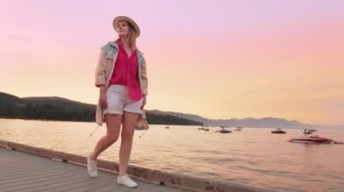 Göl kenarındaki şık trençkotlu gezgin canlı pembe ve mor günbatımı gökyüzü