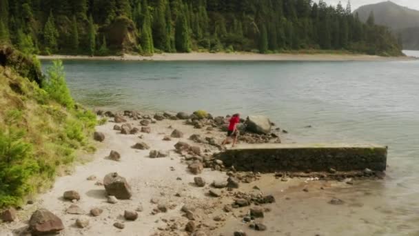 Hombre, vistiendo camiseta roja observa hermoso lago con montañas volcánicas detrás — Vídeos de Stock