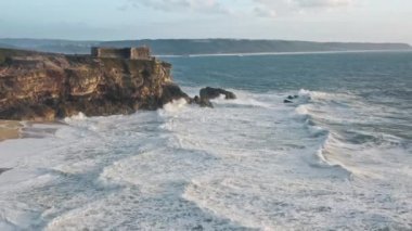 Vahşi dalgalar kıyı şeridine çarpıyor ve arka planda güneş doğuyor.