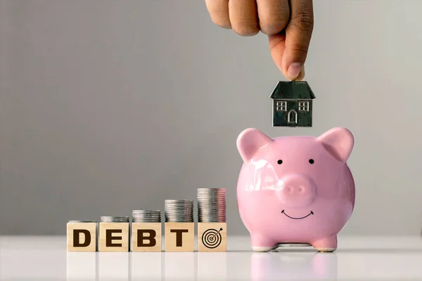 债务和金融目标的概念 在贴有债务标签的堆积如山的木块上的硬币和粉红猪储蓄中的手持房屋模型 — 图库照片