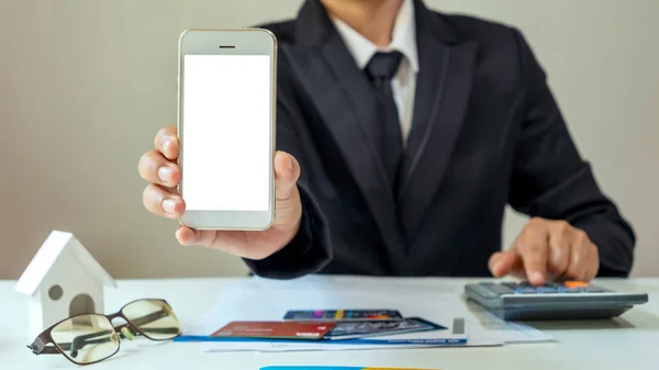 businessman holding a cell phone with a white screen and financial reports on his desk
