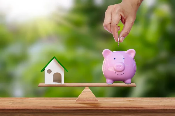 スケール上に配置された豚の貯金箱にコインを保持するモデルハウスと手とぼやけた緑の背景 不動産のアイデアと将来の投資事業計画 — ストック写真