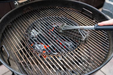 Wide angle view of a person cleaning a charcoal grill using a wire brush. Glowing coals visible beneath the grate. Outdoor barbecue maintenance scene with focus on preparation, cleanliness and grilling equipment.