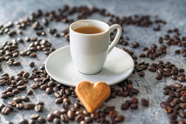 A white espresso cup filled with freshly brewed coffee stands on a saucer surrounded by roasted coffee beans. A heart shaped cookie is placed in the foreground on a grey stone surface.