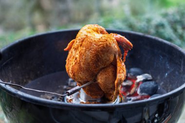 A whole seasoned chicken is roasting on a vertical poultry holder inside a charcoal kettle grill. A temperature probe is inserted to monitor the internal heat while glowing coals surround the meat.