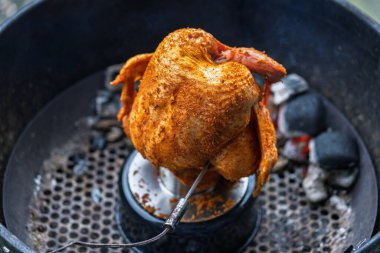 A whole raw chicken covered with dry spice rub is placed on a vertical poultry roaster inside a charcoal kettle grill. Glowing briquettes and rising heat prepare the meat for indirect grilling outdoors.