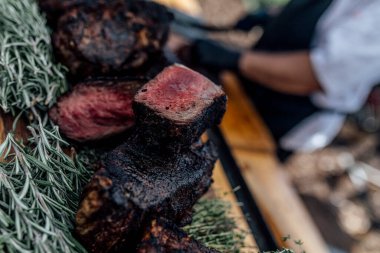Tütsülenmiş et eti bir barbekü etkinliği sırasında ahşap bir kesme tahtasıyla taze dilimleniyor. Kömürleşmiş bir kabukla taze eti yakın, kırsal ve otantik bir mutfak havası için doğal ışık altında yakalanır.