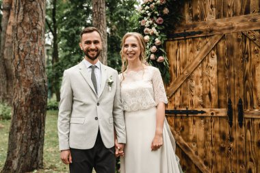 Genç çift evleniyor, açık havada nikah töreni mutlu yeni evliler el ele tutuşuyor, damadın arka planında gelin ile düğün kemeri, taze çiçekli ahşap kemer ve renkli güller.