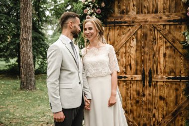 Genç çift evleniyor, açık havada nikah töreni mutlu yeni evliler el ele tutuşuyor, damadın arka planında gelin ile düğün kemeri, taze çiçekli ahşap kemer ve renkli güller.