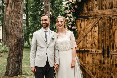 Genç çift evleniyor, açık havada nikah töreni mutlu yeni evliler el ele tutuşuyor, damadın arka planında gelin ile düğün kemeri, taze çiçekli ahşap kemer ve renkli güller.