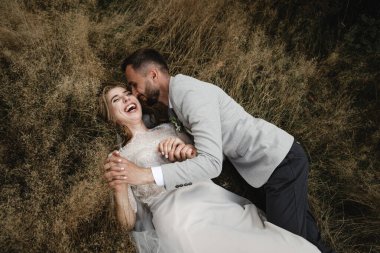 Güzel, mutlu gelin ve şık damat çimenlere uzanıp kucaklaşmanın tadını çıkarıyorlar. Düğün günlerinde mutlu gelin ve damat. Yeni evliler birlikte mutlu, harika bir düğün günü.