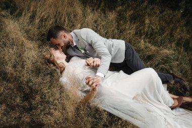 Güzel, mutlu gelin ve şık damat çimenlere uzanıp kucaklaşmanın tadını çıkarıyorlar. Düğün günlerinde mutlu gelin ve damat. Yeni evliler birlikte mutlu, harika bir düğün günü.