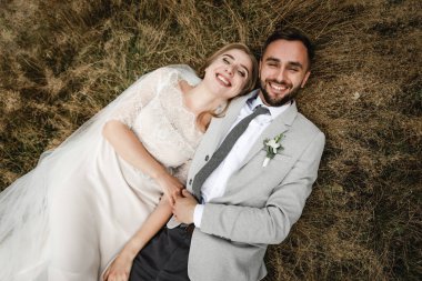 Güzel, mutlu gelin ve şık damat çimenlere uzanıp kucaklaşmanın tadını çıkarıyorlar. Düğün günlerinde mutlu gelin ve damat. Yeni evliler birlikte mutlu, harika bir düğün günü.