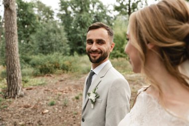 Mutlu ve neşeli gelin ve damat düğün gününde parkta yürürler ve birbirlerine bakarlar, ön planda damat arka planda mutlu bir eş, yeni evliler törene giderler.