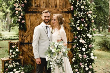 Çiçeklerle süslenmiş ahşap kemerin arkasında poz veren mutlu yeni evli bir çift. Çiçeklerle süslenmiş ahşap bir kemerin arkasında poz veren mutlu bir çift..