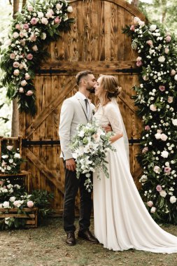 Çiçeklerle süslenmiş ahşap bir kemerin arkasında poz veren mutlu yeni evli bir çift. Çiçeklerle süslenmiş ahşap bir kemerin arkasında poz veren mutlu bir çift. Damat, elinde lüks bir buket çiçek tutan geline nazikçe sarılıyor.