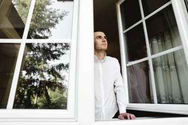 Beyaz gömlekli genç adam evde düğmesini ilikliyor. Damat toplanıyor. Damat evde. Adam giyiniyor. Kendini izole eden adam pencereden bakar.