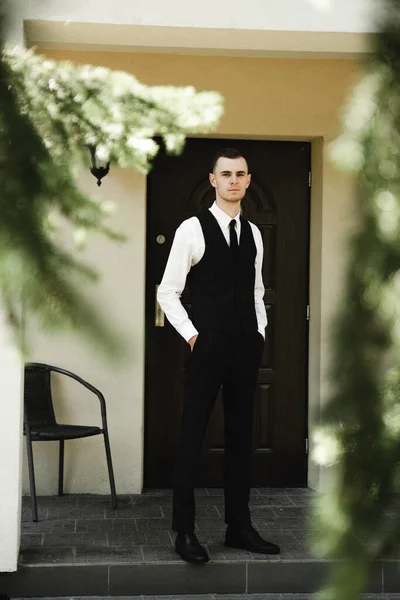 a guy dressed in suit pants and a shirt on top of which a classic vest stands near his house. the Butler. house attendants. stylish clothes of the young groom