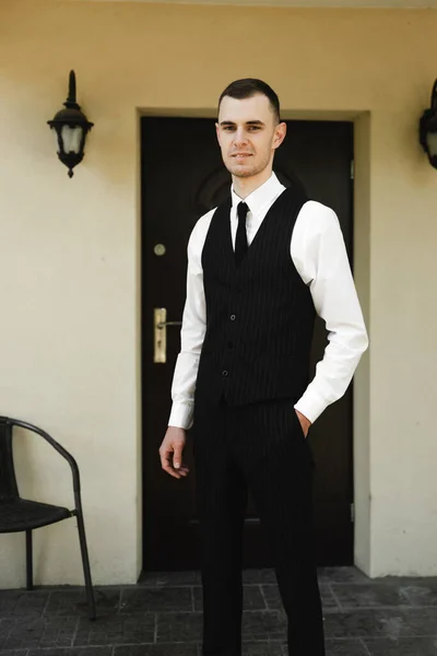 a guy dressed in suit pants and a shirt on top of which a classic vest stands near his house. the Butler. house attendants. stylish clothes of the young groom