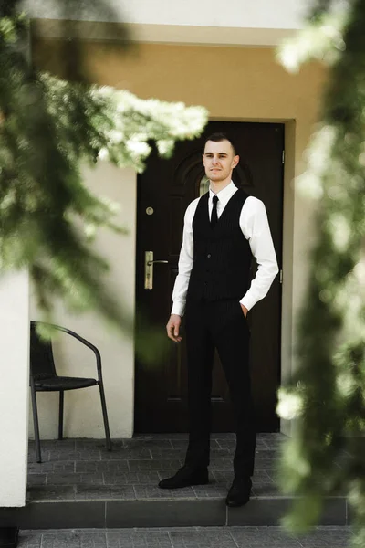 a guy dressed in suit pants and a shirt on top of which a classic vest stands near his house. the Butler. house attendants. stylish clothes of the young groom