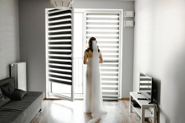 the bride is waiting for the groom in a room with blinds hanging on the window. girl at the window. silhouette of the bride. shutters on the windows. wedding day fees
