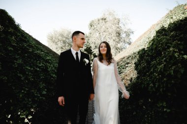 Zengin damat ve gelin, sarmaşıkla kaplı parkta güzel güzel yürüyorlar. Genç bir ailenin düğün günü. Yeni evliler. Evliliğin başlangıcı. Güçlü bir aile. romantik ilişki