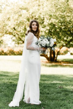 Kar beyazı elbiseli güzel gelin gün batımında elinde bir düğün buketi tutuyor. Mütevazı ve tatlı gelin. Gelinin güzel bir gelinlik içindeki portresi.