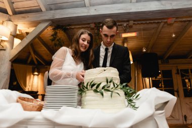 Damat ve gelin düğün pastasını romantik bir ortamda kestiler. Beyaz düğün pastası masanın üzerinde duruyor. Yeni evliler bir ziyafette pasta kesiyorlar. Çiçekçilikle süslenmiş tatlı düğün pastası