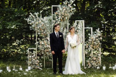 Yeni evlilerin ayakta durduğu düğün töreni. Damat ve gelin düğün sırasında sokaktalar. Çiçeklerle süslenmiş bir düğün kemerinin arka planında yeni evliler. Açık hava düğünü