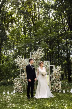 Yeni evlilerin ayakta durduğu düğün töreni. Damat ve gelin düğün sırasında sokaktalar. Çiçeklerle süslenmiş bir düğün kemerinin arka planında yeni evliler. Açık hava düğünü
