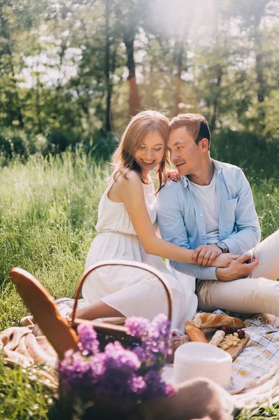 愛の素敵なカップルは ベッドスプレッド上の花や食べ物と公園のウィッカーバスケットでピクニックを組織しました 幸せな恋人は笑ってピクニックで食べる ロマンチックなデート — ストック写真