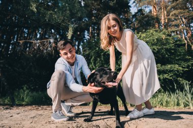 Güzel bir çift tasmalı siyah bir labrodor ile yürüyor. Sevgililer gülümser ve köpeğe bakar. Safkan bir köpekle parkta yürüyüş yapmak. Aşıkların eğlencesi. Genç mutlu insanlar.