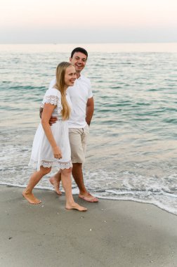 Okyanus kıyısında sahilde yürürken bir erkekle kucaklaşan mutlu bir kız. Romantik yürüyüş. Karı koca deniz kenarında gülüp sarılıyor. Yaz tatili. Genç bir çiftin tatili.