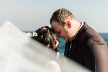 Damat ve gelin kafalarını denizin arka planına bastırdılar. Kızıl sakallı, kahverengi elbiseli tombul bir damat geline sarılıyor. Düğün fotoğrafı çekimi. genç aile
