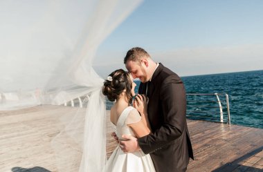Güzel bir çift gelin ve damat, iskelede, denizin arka planında ve su sıçratan dalgalarda birbirlerine sarılıyorlar. Çift deniz kenarında yürür. Denizde mutlu yeni evliler. Şirin bir çift.